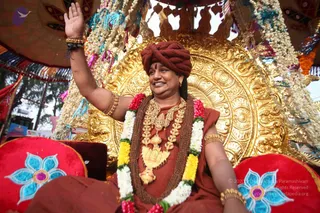 A seated figure in a procession vehicle raises his right hand in greeting while smiling. He wears a maroon-red turban, a rust-orange robe, a rudraksha mala with a large ornate gold necklace and pendant, and a garland of white jasmine, yellow and pink flowers. Behind him is a gold-embossed backrest on a golden throne-like seat, decorated with white flower strands and blue-teal tassels, with a red cushion bearing blue flower embroidery at the lower left. A red-and-gold canopy frames the scene. a person appearance cues include the saffron-orange robes, turban and ornate gold necklaces; he is seated on a ceremonial throne as a procession figure.