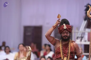 A bearded man with a dark hair bun wrapped in a rust-red turban with a jeweled ornament stands on a stage holding up a small golden leaf-shaped object in his raised right hand. He wears a rust-red shawl over his bare shoulder, a heavy gold necklace, layered rudraksha malas and red wrist threads. Beside him is a golden trident-tipped staff and a polished brass vessel shaped like a swan or bird. Blurred seated devotees in white and pastel saris fill the background of a lavender-draped hall with a wooden door.