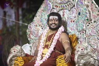 A bearded man with long swept-back dark hair sits smiling on an ornate silver throne encrusted with red, green and blue gemstones and peacock motifs, its armrests carved as elephant heads. He wears a rust-red wrap, a thick gold necklace, a gold pendant, and a pink and white flower garland, with gold bangles on his wrists and upper arm. A white horizontal tilak with a red bindi marks his forehead. Striped orange-and-black tiger skins cover the seat and footrest. The page identifies this as Swamiji, appearance-based label: Swamiji (probable), seated on a throne, with long hair, gold ornaments, rust-red clothing and garland cues consistent with the page. Background shows a greenery and a white fan.