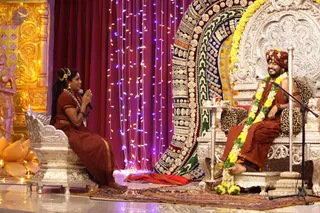 A photograph of a stage event at a temple. A woman in a rust-red sari with hair in a bun and gold hair ornaments sits on a silver chair at left, palms joined in greeting and holding a microphone. A man in an orange-red turban, orange-red dhoti-style garment, yellow-and-green garland and bare torso sits on an ornate silver throne at right, leopard-print cushions behind him, a microphone stand beside him and a small table with a glass. Behind them hang a magenta curtain with fairy lights and a large embroidered panel of gold and multicoloured motifs. A golden carved wall panel and a gold lotus decoration appear at left. The man has distinctive turban, bare chest and garland cues; the page indicates a person was present, so the appearance-based label applies.