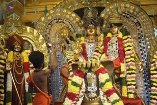 Photograph inside a gold-decorated temple shrine showing a priest in an orange dhoti with bare upper torso and rudraksha mala, raising an arm with a small object toward ceremonial figures. Behind him stand large gold and jewelled deity murti, one with a tall ornate crown and one wearing a gold crown and necklaces, both draped in garlands of yellow, red and white flowers. A flanking figure on the left wears an orange turban, orange cloth and a gold necklace, holding a trident. A silver shivalinga with flower garlands sits in the foreground. Mirror-work panels and hanging paper decorations frame the scene.