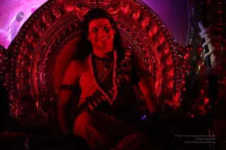 A photograph of a smiling man seated on an ornate, highly carved red-lit throne with curled scrollwork and a rounded back. He has long dark hair swept back, wears rudraksha malas and ceremonial beaded necklaces, a bare-shouldered wrap, and beaded armlets. Red and magenta stage lighting fills the scene. Swamiji (probable) appears to be the seated figure on the throne, identified by long swept-back hair, rudraksha malas, ceremonial necklaces and a bare-shouldered wrap.