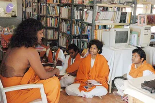 A photograph inside a library-like room lined with metal shelves of books, with a desktop computer monitor and tower on a white-covered table at right. A bare-chested man with long, swept-back dark hair sits on a white plastic chair at left, wearing an orange wrap, and speaks toward a group. Four people in orange shawls and white clothes sit on the floor: a young man in the centre holds papers and a small book, another sits near a microphone stand, and a man at right in an orange shawl looks on. A small framed picture hangs on the shelves at upper right. The left figure has distinctive long hair and a bare chest with orange wrap, consistent with a person appearance, seated as a teacher addressing students.