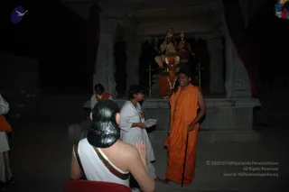 A night photograph at a temple-style shrine lit dimly, with a stone structure and a deity image of a seated pair of goddesses with crowns behind a red-draped altar bearing a garland and brass lamps. In front, a man with long dark swept-back hair wears an orange-saffron robe and holds a walking cane, standing to the right of a woman in white clothes holding a small white box. Behind them, a man in orange clothing stands near the shrine. In the lower left foreground is a white-clad figure with long black hair and gold-trimmed shawl, hands in anjali pose, seen from behind. Dark background with red curtains.