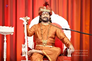 A seated man on an ornate white and red throne delivers a discourse before a red-orange drape. He wears a red-and-gold turban with a gold ornament and red feather, an orange silk kurta with a gold waist belt, a gold pendant, and a long rudraksha mala. A microphone stand is at right and a white carved reading stand at left; he holds a white ceremonial staff and smiles with eyes closed.