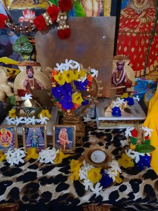 A decorated home shrine photographed at close range, with a brass deity murti at centre garlanded with white jasmine, yellow and blue flowers and orange marigold strands. Two framed portrait cards flank the central altar, each showing a bearded man in saffron robes, a red-and-gold turban and a rudraksha mala; their faces are unclear in the image. Small framed prints stand in front, including a deity in a temple gateway and a figure in a red-orange robe seated on rock. Flower-strewn brass trays hold an unlit oil lamp and small gold pyramid-shaped objects, all set on a leopard-print cloth beside a yellow garment. Rudraksha beads hang at the top left among red pompom garlands.