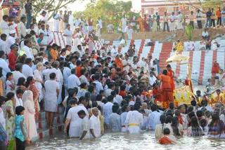 A large crowd gathers on striped red-and-white stone steps beside water, with many people standing waist-deep in the shallows. Several men in white dhotis and shirts and women in saris stand together, while a figure in orange robes stands near a golden palanquin with a canopy and marigold garlands at the right edge. Others in orange and saffron clothing move through the crowd near the palanquin, and bunting flags with paper decorations hang above the steps. Many people in the water and on the steps raise their hands or wade toward the ceremony.