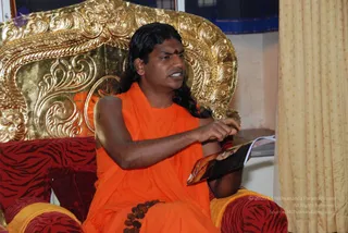 A photograph of a single man seated on an ornate gold throne with carved floral scrollwork and red tiger-striped cushions. He wears an orange saffron robe, a small red tilak on his forehead, and a rudraksha mala at his waist, with long dark swept-back hair. He holds an open booklet or magazine in his hands and appears to be speaking. A gold and yellow curtain hangs at the right edge of the room. Swamiji (probable), seated on a throne in saffron robes with long hair and rudraksha mala, appears to be giving a talk or reading.