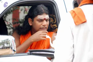 A man with long swept-back dark hair sits in the rear seat of a white car, his window open. He wears an orange saffron garment draped over his shoulders and a red bindi with white marks on his forehead. He rests his hand near his chin and looks downward while speaking. In the foreground, a person in white clothes with an orange shawl stands at the window. A palm tree and a red building are visible through the car window and side mirror. Printed text is faintly visible on the white clothing, unreadable. Swamiji (probable): the seated man shows long swept-back hair, saffron clothing and forehead markings.