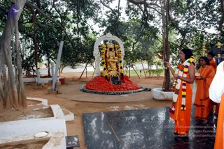 Outdoor photo beneath large banyan trees at an ashram shrine. A golden deity murti seated on a red-draped platform is garlanded with multicolour flower garlands and set within a white arched frame. A man in saffron/orange robes with long dark hair, a white floral garland and beaded necklaces holds a lit oil lamp or aarti flame toward the murti. Beside him, another saffron-robed person with a beaded necklace holds offerings. A camera operator is visible at the right edge, and a white-clad figure stands at the far right. The ground has white kolam-style patterns and a dark stone slab in front of the shrine.