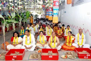 A photograph of a homa or yagna ceremony in a decorated hall, with a row of seated devotees in front of red fire-pit trays holding ash, wood and offerings. In the foreground, a bald man in a white dhoti and a bare-chested figure with a rudraksha-style necklace sit cross-legged; a woman in an orange sari sits beside them. Most participants wear white dhotis and shirts, with yellow marigold garlands. Paper lanterns and hanging decorations fill the ceiling, and a row of people in white sits behind in the background.