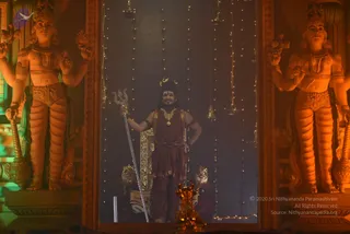 A standing man in a maroon-red sadhu-style robe with a gold necklace and rudraksha malas holds a silver trident and poses before a dimly lit shrine. Two large orange-lit guardian statues flank him, each with multiple arms, jewelled armlets and a crown, and a brass oil lamp sits in the foreground beside strings of fairy lights and hanging bells. He has long dark hair and a tilak, and the figure stands in front of a gold-trimmed throne with a tiger-skin design visible behind him. The setting looks like a darshan stage in a Hindu temple-style hall.