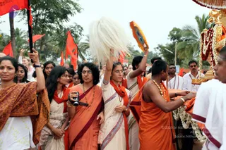 Outdoor procession with devotees following a decorated golden ratha or chariot on a palm-lined road. A woman in cream sari with red-and-maroon border raises a white yak-hair chamara fan overhead. Beside her, a woman in orange sari holds a camera, and a woman in orange-and-red garments holds a folded orange cloth. A man in orange dhoti with bare chest and gold chain necklaces stands near the chariot's garland-decorated side, holding marigold garlands. Red flags on poles rise behind the crowd. Several women wear saffron and red saris, and a man in white stands at the right edge beside the chariot with flowers and gold fittings.