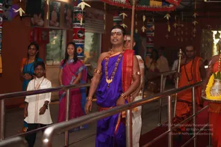 A temple interior during a procession or pada-puja. A man in a purple dhoti with a red-and-gold border and a gold-trimmed red shawl walks forward, wearing a gold necklace, a long rudraksha-style garland, gold armbands and a red tilak on his forehead. Behind him a man holds a tall staff under a red-and-gold canopy, and a saffron-robed priest carries a silver tray of yellow flower garlands. A woman in a pink sari and another in blue stand with hands joined, and a boy in white kurta watches from the left. Metal railings line the red-carpeted walkway and colourful hanging banners decorate the walls.