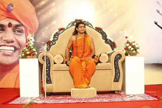A man seated on an ornate beige throne-style armchair on a red carpeted stage, with a large backdrop portrait behind him of a smiling man wearing an orange turban and red tilak. The seated man wears an orange saffron robe, a long rudraksha mala and a red bindi, with long swept-back hair and bare arms, and rests his hands on the armrests. Red and white floral arrangements flank the chair, and a microphone stand stands at the left edge. The backdrop shows raised hands at the right edge. A ceremonial discourse setting.