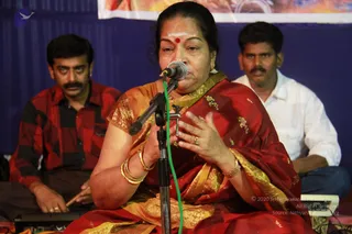 A woman in a red and gold silk saree sings or speaks into a microphone on a stand at a stage performance. She wears a red bindi and vertical vibhuti-style marks on her forehead, gold jhumka earrings, gold bangles and a gold wristwatch, and gestures with her right hand. Behind her, two men sit on the left and right: one in a red-and-black checked shirt playing a percussion instrument, the other in a white shirt. A blue backdrop and a partly visible banner appear behind the stage, with a green microphone cable running down the stand.