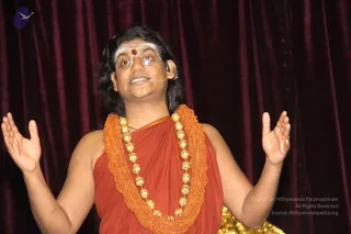 A man stands on a stage before a maroon velvet curtain, arms raised with palms open as he speaks. He wears a rust-red saffron-toned shawl draped over bare shoulders, a heavy orange rudraksha mala with gold beads, and a white forehead band with a red bindi. A small headset microphone is at his ear, and a gold drape is visible at the bottom edge. Appearance-based label: Swamiji (probable), seen in person at a discourse, with long swept-back hair and rudraksha and gold ornaments as qualifying cues.
