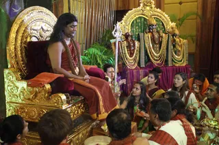 A seated man with long dark hair wears a maroon-and-rust robe and a beaded mala with a gold pendant, sitting on an ornate gold throne with a maroon cushion and an orange cloth draped over its arm. Before him, a row of devotees in saffron, maroon and green saris and kurtas gather closely; a woman in green holds a small brass vessel and others hold cups and ritual items. Behind them stands a gold-framed shrine with two gold-crowned deity murti adorned with white and gold garlands on a purple draped altar, and a silver trident stands upright in the foreground. Green foliage and a wood-panelled wall complete the setting. a person is seated on the throne, identified by his long swept-back hair and maroon-and-saffron robe with a gold-beaded mala.
