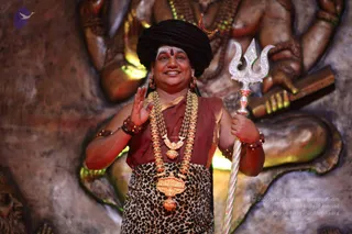 A photo of a smiling man standing in front of a large carved stone backdrop of a multi-armed deity with a crescent ornament visible above. He wears a black turban, a brown-red top with gold trim, a leopard-print lower garment, and a large gold necklace with ornate pendants and beaded mala strands; he has a red bindi and white forehead markings. He raises one hand in greeting and holds a silver trishul in the other, with red beaded bracelets on his wrists. a person. One person is visible.