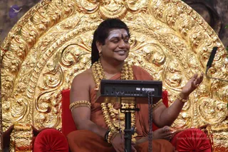 A seated man in saffron-orange robes sits on an ornate gold throne with red cushions, smiling with eyes closed and one hand raised in an open gesture. He wears layered gold necklaces, gold bangles and a gold armband, with a tripundra mark of white and vibhuti on his forehead and a kumkum dot above the brow. A black Yamaha keyboard unit sits on a stand in front of him. Based on the page's identification and the distinctive saffron robes, rudraksha-style gold beads, ceremonial gold necklaces and ornate throne, this is a person, seated and giving a discourse.