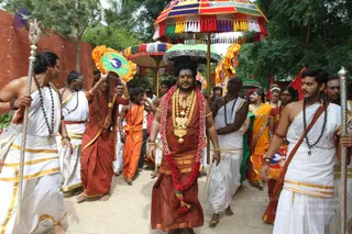 A procession on a paved path with trees behind. A central figure with long dark hair, wearing a red-brown dhoti and shawl, a red and white flower garland, a large gold necklace and gold armbands, walks forward at the center. Around him, men in white dhotis with gold borders and rudraksha malas carry a silver trident-like staff and walk barefoot. Behind them, a colourful striped ceremonial umbrella and a green-and-gold umbrella are held aloft, and attendants in orange and saffron cloth carry a fan with a green and orange border. Women in orange and yellow saris and gold ornaments walk in the procession. A red wall stands at left. The scene resembles a ritual procession, with visible ceremonial umbrellas, staves and fans.