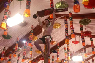 A shirtless man with a black headband and dark shorts balances on top of a tall wooden pole, holding a second person horizontally above his head with both arms extended. The supported person wears a red garment and is stretched out face-down with arms and legs hanging. The ceiling above is hung with colourful cylindrical banners in orange, yellow, red and green, decorated with birds and floral motifs, plus round lanterns and tassel umbrellas. Bright white lights illuminate the hall.