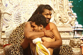 A man with long swept-back dark hair, a white forehead band, and a rust-red cloth draped over his shoulders embraces a younger man from behind while seated on an ornate throne. The throne is covered with a leopard-print cushion and silver-embossed carvings, and carved golden lion figures sit at its arms. The younger man, bare-chested with a white and yellow sash across his waist, leans into the hug and smiles. Behind them, a green backdrop and painted temple-style tower are visible.