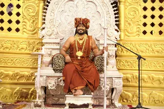 A smiling man with a full beard sits on an ornate silver throne with leopard-print cushions and carved lion armrests, set against gold-carved walls with elephant and horse reliefs. He wears a saffron-orange turban, orange shawl and dhoti, multiple layered gold necklaces and a large gold pendant, with orange rudraksha-style beads and gold bangles on his arms, and holds a tall silver staff. A small framed portrait sits on a side table, and a microphone stand stands to the right. Swamiji (probable): the saffron turban, layered gold necklaces and ceremonial throne match the page's claim, so this is treated as an appearance-based candidate seated in person, with the sitter presented as a seated teacher during a satsang.