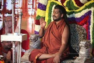 A seated man in an orange-saffron robe with long dark hair and a tilak on his forehead raises his right hand in a gesture of blessing while smiling. He sits on a leopard-print cushioned throne flanked by two silver lion figures, in front of a backdrop of garlands in yellow, white, purple and red. To the left, an elderly man with glasses and a red shawl sits with palms pressed together in anjali, and another man in glasses with a red shawl sits with folded hands near a silver stand holding a small cup. The setting resembles a satsang or public address hall with ornate red and gold decor.