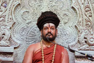 A seated man in a large dark dreadlocked turban wrapped with rudraksha beads, a red tilak on his forehead, and a dark beard, wearing a reddish-orange shawl draped over his bare shoulders and multiple heavy gold bead malas. He sits before an ornate silver repoussé backdrop with floral and scrollwork patterns, a microphone on a stand near him. Swamiji (probable): the turban, rudraksha, saffron-toned shawl and ceremonial gold malas match the presentation of the figure, seated in the central place of honour as a speaker.