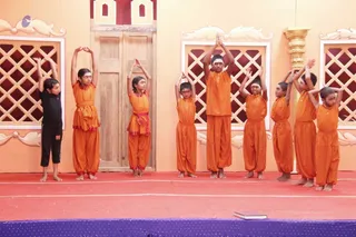 A group of nine children stands in a row on a red-floored stage with a peach-coloured backdrop featuring carved wooden doors and lattice windows. Most wear matching orange kurta-and-pant outfits with orange sashes, and they raise their arms overhead with palms pressed together in a synchronized dance or devotional pose. One child at the left wears black pants and a black top, and a closed book lies on the stage floor at the front. Two children on the right have their hands raised in a similar gesture.
