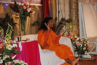A photograph of a seated figure on a white-draped couch on a stage, speaking and gesturing with one raised hand while holding a small book or paper. The figure has long dark hair and wears an orange saffron robe. Behind the couch are peacock feathers and tall bouquets of pink, red, white and yellow flowers on a pink-draped table and stands, with gold drapery and a banner in the background. The figure looks like a probable Swamiji (probable) seated in a teaching or discourse setting, with long swept-back hair and orange saffron clothing as the distinguishing cues. A small brass lamp and a bottle sit on the pink table beside the flowers.