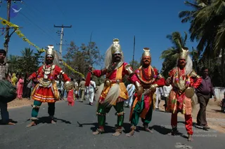 Four performers in elaborate folk costumes stand on a paved road lined with palm trees under a clear blue sky. Each wears a tall gold crown, a white feathered mane, face paint, layered beaded necklaces and gold arm ornaments; their outfits mix red, orange, green and yellow fabrics, and they hold round metal shields. Spectators in white and pastel clothing stand behind them along a dusty roadside beneath power lines and yellow streamers. The scene resembles a street procession. No a person candidate is clearly visible in the frame.