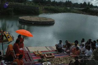 Outdoor ritual by the edge of a pond at dusk, with a circular stone platform in the water behind. Several people in orange or saffron robes sit and stand on striped woven mats, two of them beneath a bright orange umbrella and a decorated multicoloured parasol. Seated devotees in white and light clothing face the ritual, while a brass tray holds a teapot-like vessel and a flower-garlanded box. Rows of small brass lamps and offering plates are arranged on the mat in front, with fruit and a cloth bundle nearby. Overcast sky and palm trees frame the scene.