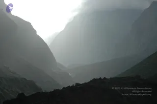 A misty Himalayan mountain landscape photographed in dim light. Steep, dark rocky ridges rise on both sides of a deep valley, with cloud and haze drifting between the peaks. A narrow winding road or track is visible along the lower slope in the foreground. No people are visible.