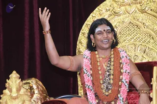 A photograph of a man seated on an ornate gold throne against a deep red velvet backdrop, raising his right hand in a gesture while speaking to an audience. He wears a saffron-orange garment, a heavy rudraksha mala with gold beads and a pink-and-white flower garland, gold earrings, and a white forehead mark with a red bindi. A gold lion-like figure and a gold bull statue sit at the lower left.