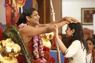 A seated man in a red-orange robe with a pink rose garland and rudraksha mala sits on a throne-like seat beside a gold peacock-feather fan and a gold kalasha. He smiles as he places both hands on the head of a woman in a white top with orange-red shawl, gold bangles and drop earrings, who gently raises her palms toward him. A silver staff stands between them. A second woman with a sandalwood-marked forehead and beads watches at right, and a framed artwork hangs on the pale wall behind. The seated man with long swept-back hair and distinctive ornate robes and garland is a person, shown as an in-person satsang blessing.