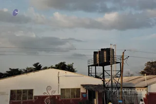 A photograph of a rural building complex under a cloudy evening sky. A white-washed building with red painted border and a decorative curling motif sits on the left, with a thatched awning and a pale-blue sign. Behind it stands a metal water tank stand holding two large black water tanks, with a corrugated metal roof and a wooden fence to the right. A small figure in white stands at the lower right near the fence, and another person in dark clothing is partly visible at the bottom centre. Power lines cross the sky, and a faint purple bird logo appears in the upper left.