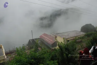 A misty hillside in the Himalayan region, with low clouds wrapping the slopes behind a cluster of buildings. In the foreground, green shrubs and a partially visible person in red headwear and a white shirt with a dark vest stand at the edge of the frame. A low concrete building with a rusty red corrugated metal roof, painted with Devanagari-style lettering, sits beside a larger yellow-walled building with a flat roof. Power lines cross the sky and a tall utility pole stands near the buildings. The scene suggests a mountain road journey in clouds.