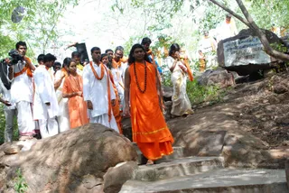 A procession of devotees walks up a stone staircase through a wooded hillside. In the centre, a young man with long dark hair wears a saffron-orange robe with a gold trim and a rudraksha mala, and walks ahead of the group. Behind him, men in white dhotis and orange shawls and a woman in an orange sari carry a flat object. To the right, a woman in a white and gold sari stands with hands joined, and a boulder bears a sign reading 'Fire is Strictly Prohibited'. Swamiji (probable): the central figure has long swept-back hair, saffron robes and a rudraksha mala, and is leading the procession.