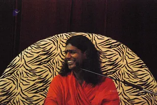 A photograph of a smiling man with long, swept-back dark hair and a dark red shawl-like wrap, seated in front of a large cream-and-black tiger-stripe patterned backdrop on a dark red curtain. A microphone stand is visible at the right edge.