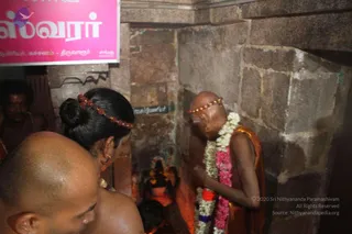 A photograph inside a dim stone shrine with rough grey granite walls. A bald, bare-chested older man in an orange-saffron cloth wraps his shoulders and wears a white and red-pink flower garland and a beaded headband; he stands with hands folded in front of a small deity murti niche. Beside him a woman with hair in a bun and red flower adornment, and a bald bare-chested man in the foreground, face the shrine. A pink sign with Tamil text hangs above, and a small lamp and offerings are visible in the niche.