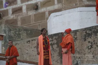 Outdoor photograph beside a weathered brick and plaster wall at a stone ghat-like setting. Three saffron-clad men stand on a ledge. On the left a man in orange shawl stands with hands clasped. In the centre a man with long dark hair wears a peach-orange shawl, a multicolour flower garland and a rudraksha-style mala over a maroon top, with a gold-striped maroon dhoti. On the right an elderly bearded sadhu in a red-orange turban and orange shawl over pale pink lower cloth raises one finger toward his face, facing the centre man.