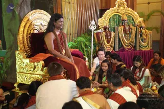 A seated man with long dark hair, wearing a rust-red robe, rudraksha-style beaded necklaces and a gold chain, sits on an ornate gold-and-crimson throne during a puja. Devotees in white and saffron-orange shawls sit on the floor before him, some offering fruit and flowers on brass plates while a woman in a green sari and another in a white outfit with red shawl assist. Behind the throne stand adorned deity murtis draped in garlands, and a silver trident is planted near the seat. Tropical leaves and bamboo decorate the stage. Probable Swamiji (probable) on the throne, with long swept-back hair, rudraksha beads and saffron-red robes.