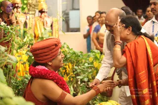 A photograph of a devotee in an orange-red turban and red flower garland kneeling and smiling while reaching toward a seated older man in white clothes with a grey shawl and a red-and-white forehead mark. An older woman in an orange sari with gold border and red bangles raises her hand to the man's face in blessing. Behind them, yellow-leafed plants surround deity murti with gold garlands, and a row of standing onlookers in white and colourful dress watches. Bananas lie at the lower right.
