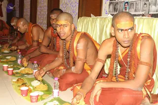 Several shaved-headed men in saffron-orange robes sit cross-legged on the floor eating from banana leaves set with rice, fried snacks and a yellow dish, with small plastic water bottles and red paper cups beside them. The men wear rudraksha malas, bangles and orange-gold sashes, and several have yellow and white sacred markings on their foreheads and arms. Behind them a pale green draped table and a wall of framed black-and-white portraits are visible.