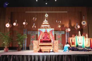 A stage at an indoor hall is arranged with a large gold-and-red throne in the centre, seated on a red cushion beneath an ornate gold canopy. A seated figure in orange-red clothing wears a pink-and-white flower garland and sits in a meditative pose with hands raised. Flanking the throne are painted blue-and-gold panels and wooden walls hung with ornamental drums and cobra motifs. To the right, a large deity murti dressed in green and gold cloth with gold ornaments stands beside a smaller figure, and a woman in a light blue sari kneels at the base of the murti. Potted palms sit on the left. The seated figure matches the appearance of a person, with the orange-red robe, garland and throne cues, and is shown on a stage.
