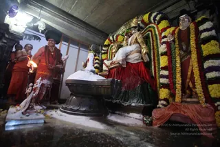 A photo of a temple shrine at night during a Mahashivaratri puja. A priest in a deep orange-red robe with a dark turban-style headwear and a rudraksha mala stands at center holding a flaming lamp, with another priest in orange beside him and devotees partly visible at left. Before them is a large white-coated Shivalinga on a dark stone yoni peetha, dusted with white ash and surrounded by a flower-strewn floor. Behind it sits a golden deity murti in a red and white garment with an ornate crown, flanked by a floral garland of yellow, white, purple and red marigolds. A decorated figure with a gold face mask and orange-red cloth stands at right, also garlanded. The scene conveys a pratyaksha padapuja ceremony at a shrine.