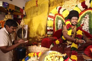 A seated man with long, swept-back dark hair sits on a red velvet throne decorated with garlands of yellow marigolds, red roses and white flowers, a golden trishul standing beside him. He wears a maroon-brown dhoti, gold necklaces, bangles and a rudraksha mala, and smiles while holding a small object. Another man in a white shirt, holding a white item, stands at left beside a dark vessel and a wicker basket. A silver plate of yellow sweets sits in front. Behind is a golden wall with carved relief. Swamiji (probable) is the seated figure on the throne, identified by his distinctive long hair, rudraksha mala, gold necklaces and ceremonial clothing.
