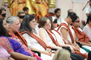 A seated group of devotees in an ashram hall with eyes covered by black blindfolds, many with eyes closed as in meditation. Several women and men wear white tops and loose white clothing with orange-red shawls bearing printed script, and some wear name badges on lanyards. A woman in the foreground wears glasses and a dark wrap; a woman on the left wears a vivid blue and pink patterned garment. Behind them, a large gold-and-orange deity murti is seated, and a microphone stand appears at the upper right. No individual is clearly identified as a person.